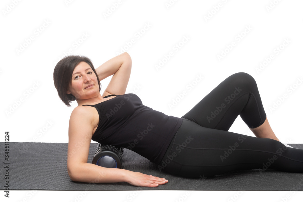A middle-aged woman with saggy skin on a gymnastic mat with myofascial roller does an exercise on her back on a white background.