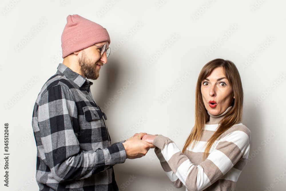 Young man with a beard in a hat and a plaid shirt and a girl in a sweater hold hands, a girl with a surprised face on an isolated light background. Emotional face. Happy couple dating Young man with a beard in a hat and a plaid shirt and a girl in a sweater hold hands, a girl with a surprised face on an isolated light background. Emotional face. Happy couple dating