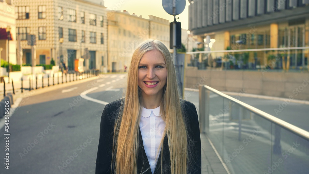 Elegant blond woman in suit standing on street with smile against urban background.