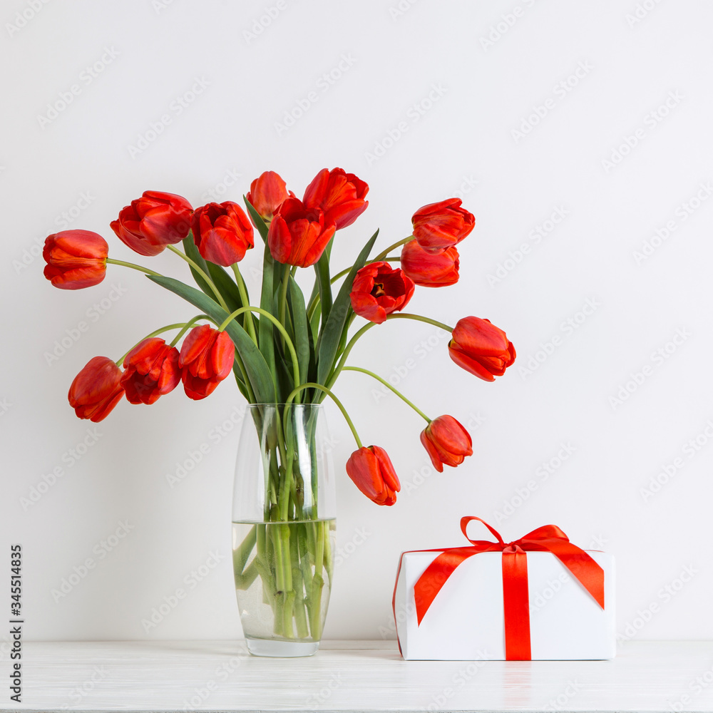 Red tulips in a vase and gift on the table, on a white background. Postcard blank.