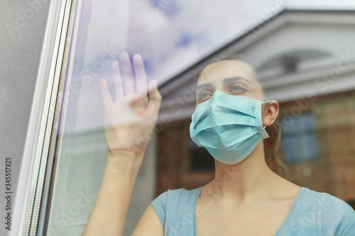 health, safety and pandemic concept - sick young woman wearing protective medical mask looking through window at home