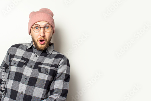 Portrait of a young man with a beard in a hat, a plaid shirt and glasses looks out from the side with a surprised face on an isolated light background. Emotional face. Gesture surprise shock