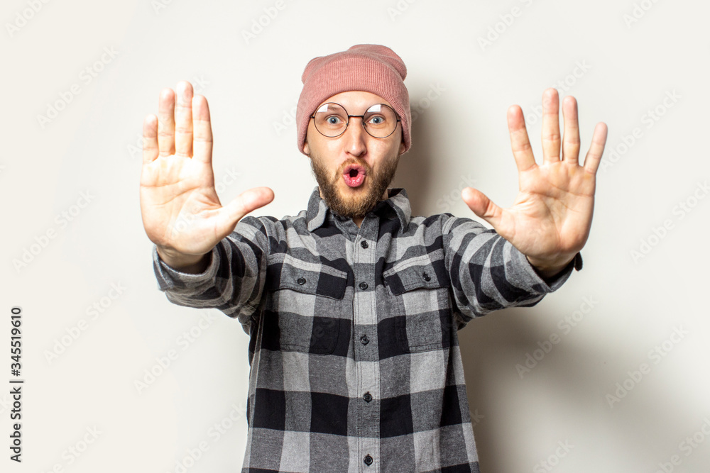 Naklejka premium Portrait of a young man with a beard in a hat, a plaid shirt stretched out his hands in front of him with his palms to the viewer on an isolated light background. Emotional face. Stop gesture, enough