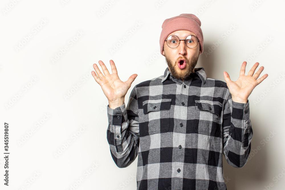 Portrait of a young man with a beard in a hat, plaid shirt and glasses with a surprised face throws up his hands on an isolated light background. Emotional face. Gesture of surprise, shock Portrait of a young man with a beard in a hat, plaid shirt and glasses with a surprised face throws up his hands on an isolated light background. Emotional face. Gesture of surprise, shock