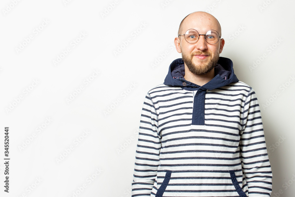 Obraz premium Portrait of a Bald Young man with a beard in glasses, a sweater with a hood, smiling on an isolated light background. Emotional face