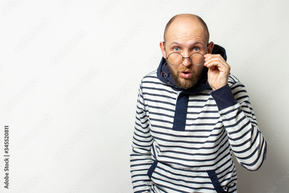 Portrait of a Bald Young man with a beard, a sweater with a hood lowered his glasses on his nose with a surprised face on an isolated light background. Emotional face. Copy space Portrait of a Bald Young man with a beard, a sweater with a hood lowered his glasses on his nose with a surprised face on an isolated light background. Emotional face. Copy space