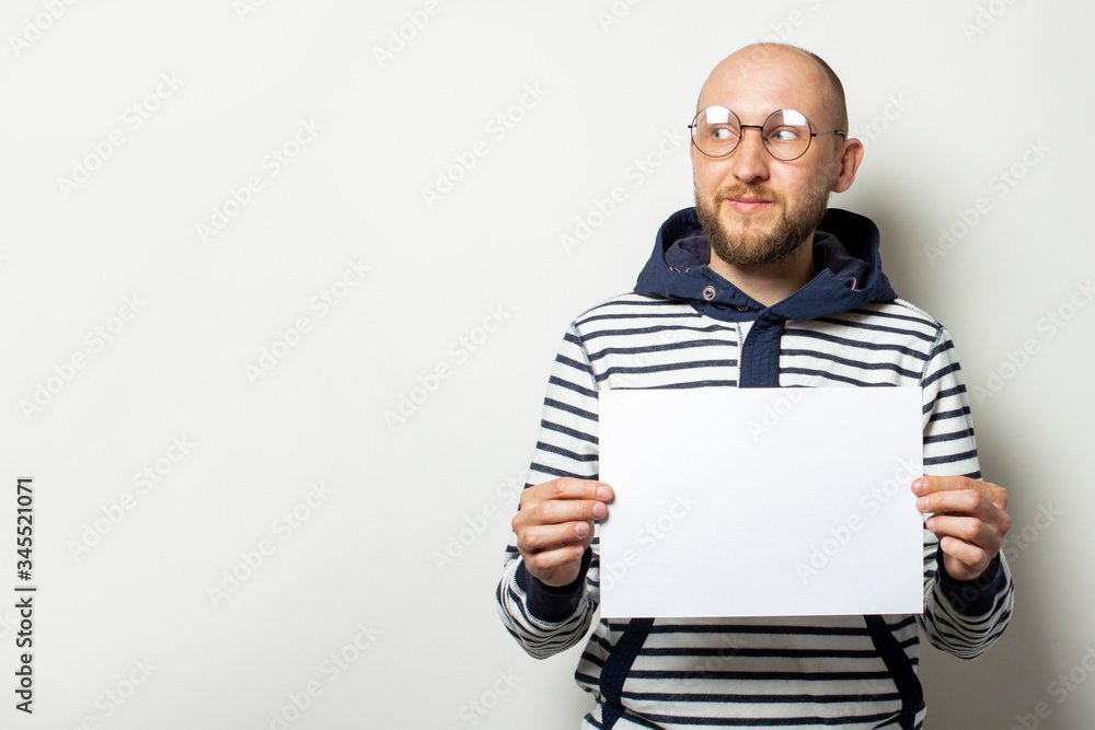 Portrait of a Bald Young man with a beard in glasses, a sweater with a hood holding a blank sheet of paper in front of him, looking to the side on an isolated light background. Copy space Portrait of a Bald Young man with a beard in glasses, a sweater with a hood holding a blank sheet of paper in front of him, looking to the side on an isolated light background. Copy space