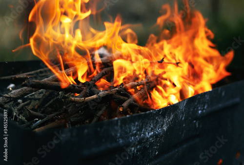 Dry, brown, small tree branches, which is in the black barbeque, burn in the bright- red fire. Fire flame is very strong and aggresive. Blurred background.