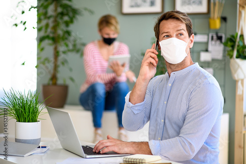 Mature businessman and colleague with face mask indoors working in office.