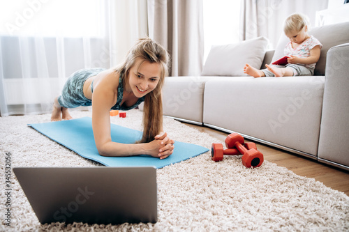 Sport at home. Young attractive mom doing sports at home and watching a video fitness lesson on a laptop, and her little baby girl sit on a couch photo