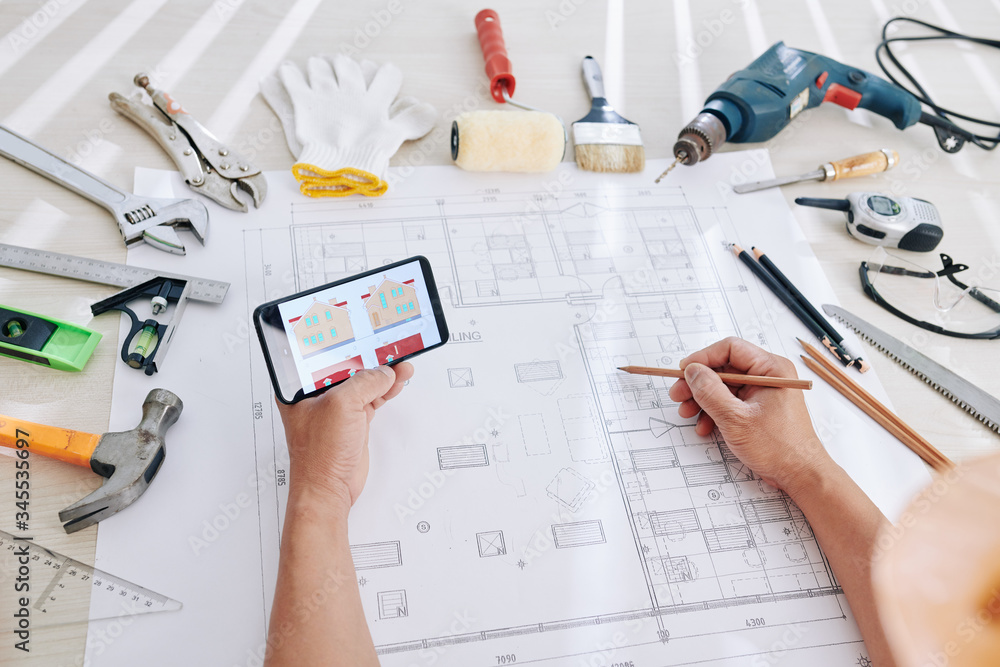 Architect checking house image on smartphone screen when working on blueprint and interior design Architect checking house image on smartphone screen when working on blueprint and interior design