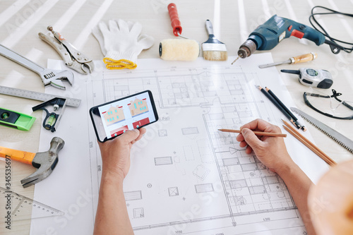 Architect checking house image on smartphone screen when working on blueprint and interior design