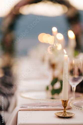 Close-up of a wedding dinner table at reception. A beautiful thin white candle burns in a small gold candlestick, frozen drops of wax dripping down. 
