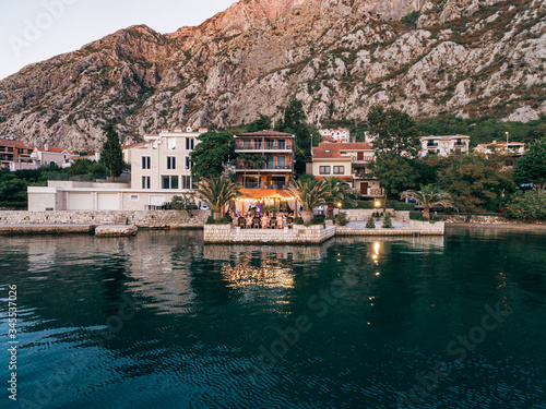 Wedding Reception Table Setting, dinner by the sea aerial. Guests are sitting at a table by the sea, amid a hotel-restaurant and large rocky mountains. Wedding in Montenegro, Kotor Bay, Dobrota