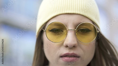 4k. Young serious girl in yellow cap and glasses stand in  city area. Close up portrait