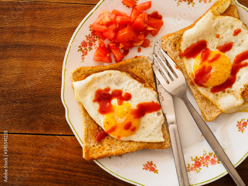 A prepared meal on an ornamental dinner plate. Consisting of, two fried eggs sunny side up, placed on two slices of toast, with some chopped tomato and a dash of ketchup, and a liver knife and fork.