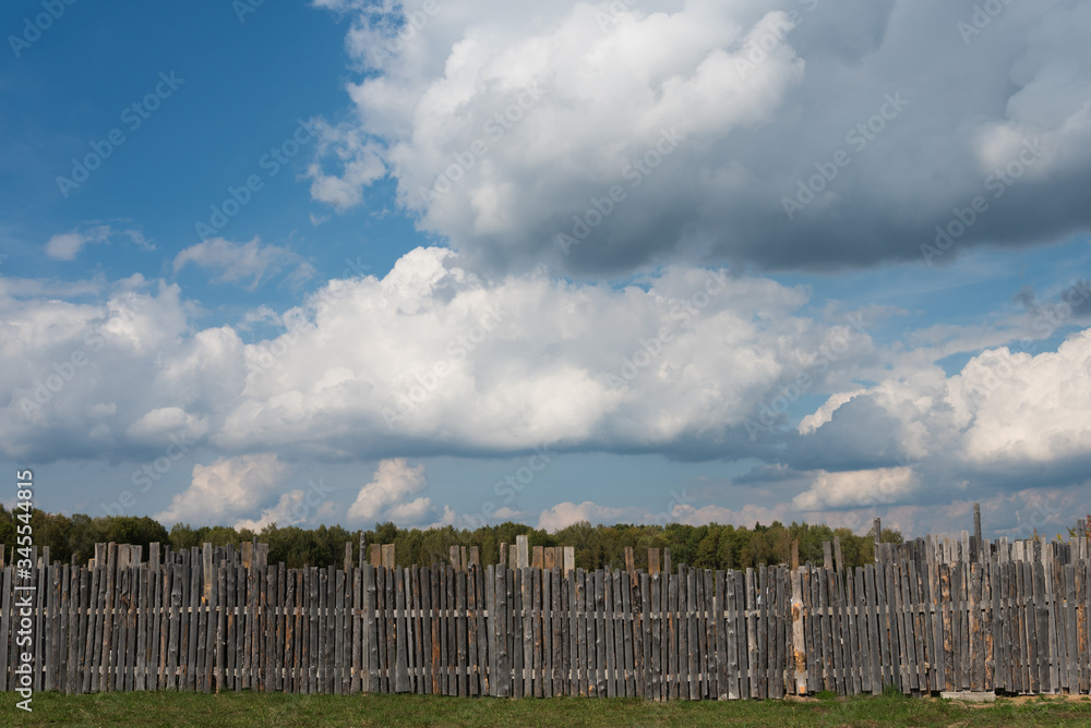 A wooden fence, behind which you can see green trees. Blue sky with elegant white clouds A wooden fence, behind which you can see green trees. Blue sky with elegant white clouds