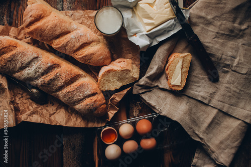 A piece of french baguette with butter on the linen fabric, craft paper and dark wooden background. Wodeen knife and eggs. One glass of milk. Craft paper photo