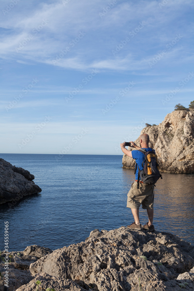 A young and bald man taking photographs with the mobile to a seascape on a beach without people in Mallorca. The man is wearing a backpack and it is early in the morning