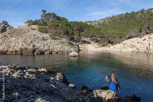 A young and bald man taking photographs with the mobile to a seascape on a beach without people in Mallorca. The man is wearing a backpack and it is early in the morning photo