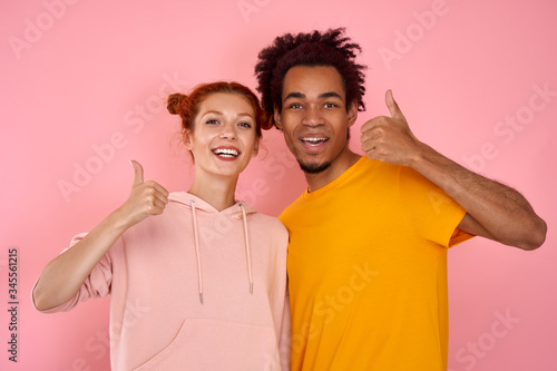 Portrait of satisfied lovely romantic multicultural couple, ginger woman and African American man shows thumb up indicate sales discounts feel excited wear modern outfit isolated over pink background.