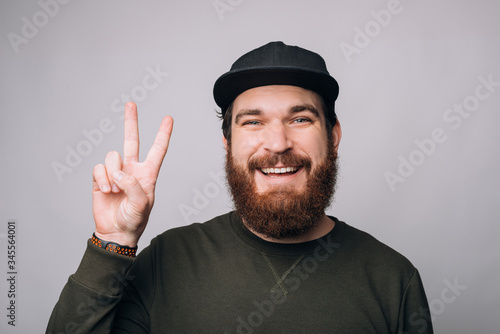 Cheerful young man wearing black cap is showing peace gesture on white background. © Vulp