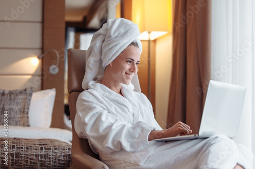 Positive successful young woman freelancer resting in a hotel room after a shower with bathrobe and towel working with a laptop while sitting at a table. Freelance success concept