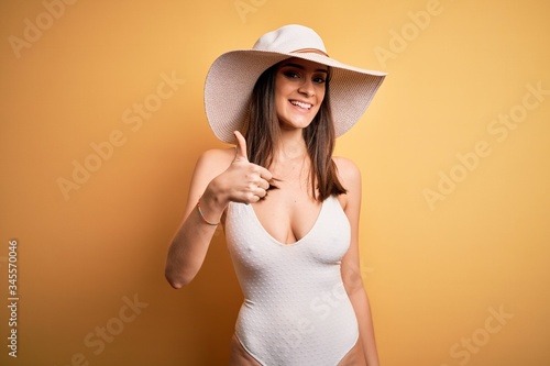 Young beautiful brunette woman on vacation wearing swimsuit and summer hat doing happy thumbs up gesture with hand. Approving expression looking at the camera showing success.