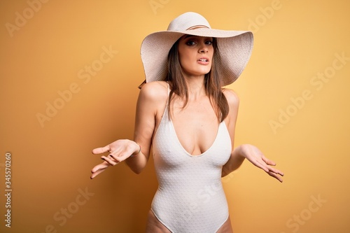 Young beautiful brunette woman on vacation wearing swimsuit and summer hat clueless and confused with open arms, no idea concept.