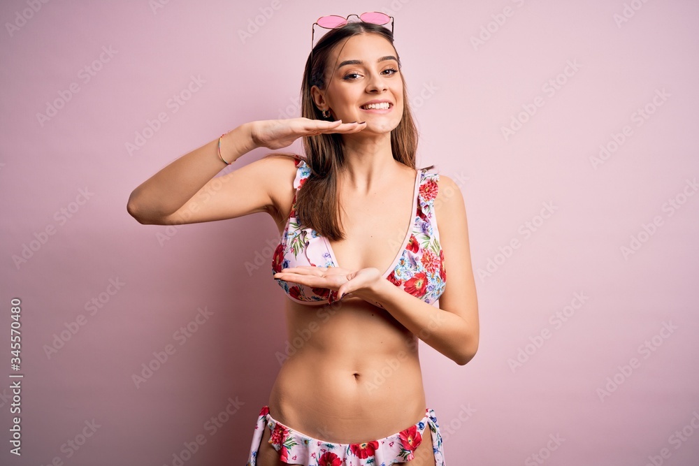 Naklejka premium Young beautiful woman on vacation wearing bikini and sunglasses over pink background gesturing with hands showing big and large size sign, measure symbol. Smiling looking at the camera. Measuring