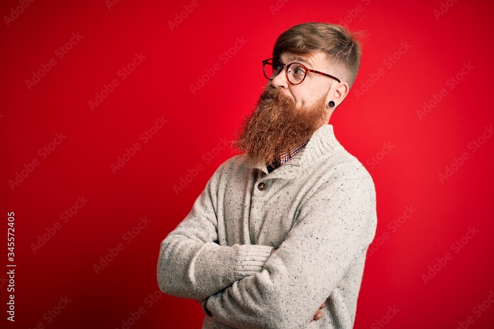 Plakat Handsome Irish redhead man with beard wearing casual sweater and glasses over red background looking to the side with arms crossed convinced and confident