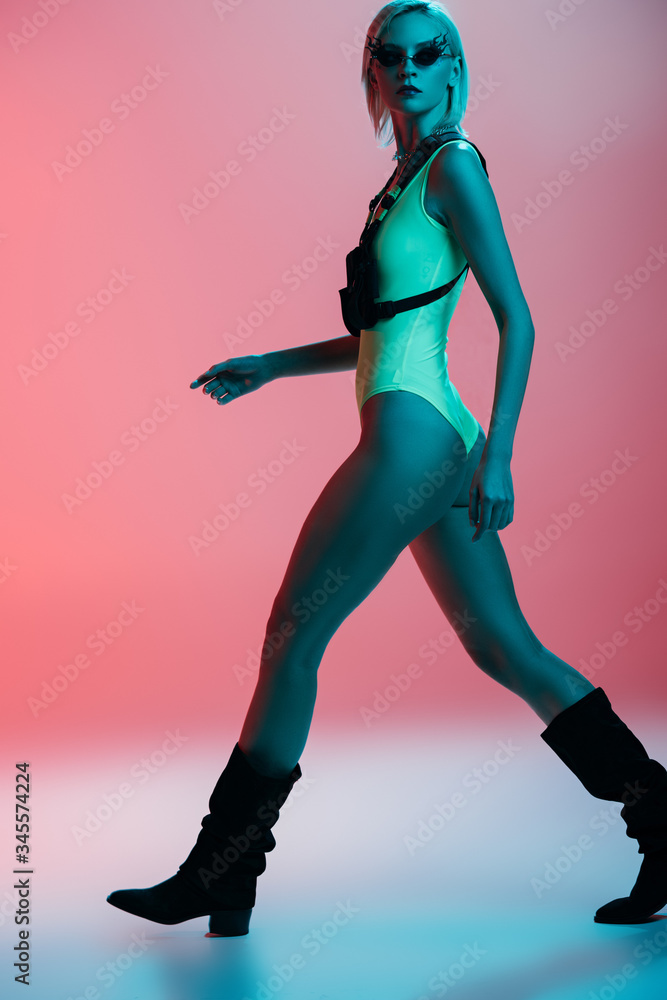 fashionable young model in futuristic leotard and fire-shaped sunglasses posing on pink in blue light