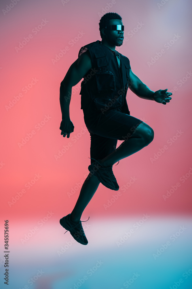 handsome fashionable african american man in futuristic sunglasses posing on pink in blue light