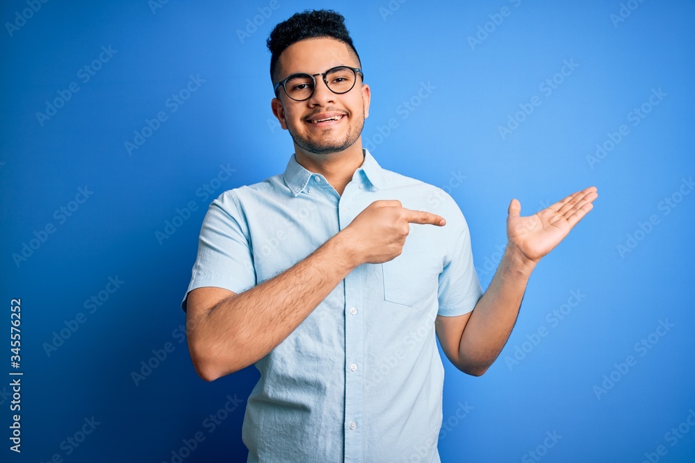 Young handsome man wearing casual summer shirt and glasses over isolated blue background amazed and smiling to the camera while presenting with hand and pointing with finger. Young handsome man wearing casual summer shirt and glasses over isolated blue background amazed and smiling to the camera while presenting with hand and pointing with finger.