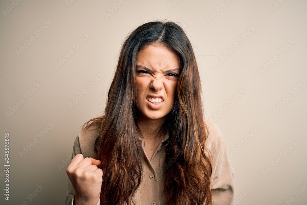 Young beautiful brunette woman wearing casual shirt standing over white background angry and mad raising fist frustrated and furious while shouting with anger. Rage and aggressive concept. Young beautiful brunette woman wearing casual shirt standing over white background angry and mad raising fist frustrated and furious while shouting with anger. Rage and aggressive concept.