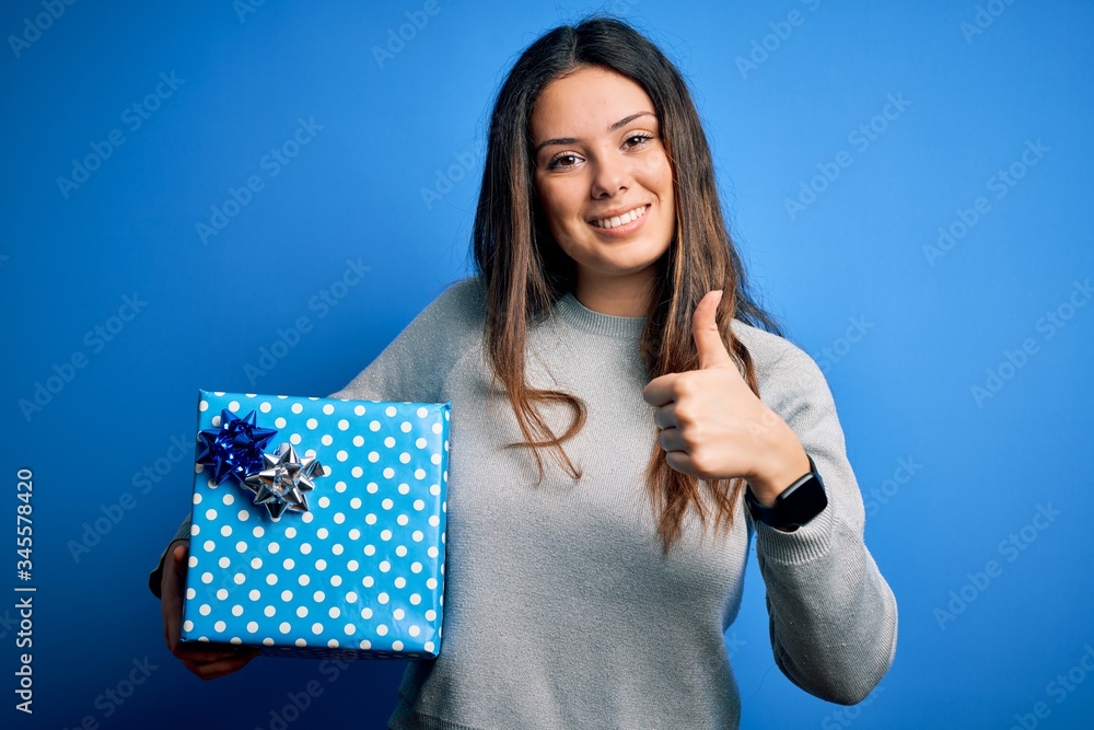 Young beautiful brunette woman holding birthday gift over isolated blue background happy with big smile doing ok sign, thumb up with fingers, excellent sign Young beautiful brunette woman holding birthday gift over isolated blue background happy with big smile doing ok sign, thumb up with fingers, excellent sign