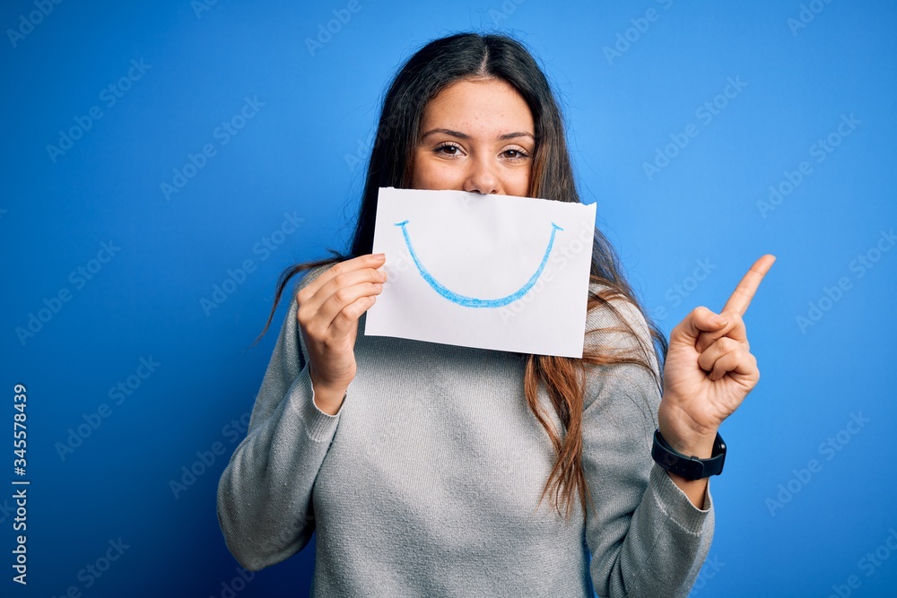 Young beautiful brunette positive woman holding card with smile emotion on mouth very happy pointing with hand and finger to the side Young beautiful brunette positive woman holding card with smile emotion on mouth very happy pointing with hand and finger to the side