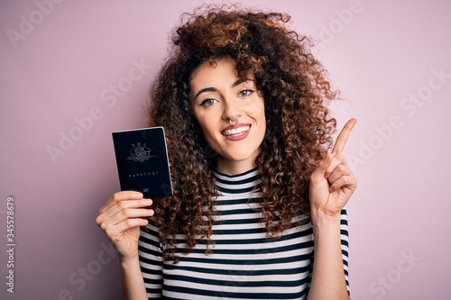 Beautiful tourist woman with curly hair and piercing holding australia australian passport id surprised with an idea or question pointing finger with happy face, number one