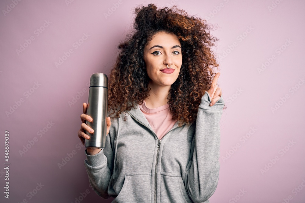 Young beautiful tourist woman with curly hair and piercing holding thermo with water surprised with an idea or question pointing finger with happy face, number one Young beautiful tourist woman with curly hair and piercing holding thermo with water surprised with an idea or question pointing finger with happy face, number one