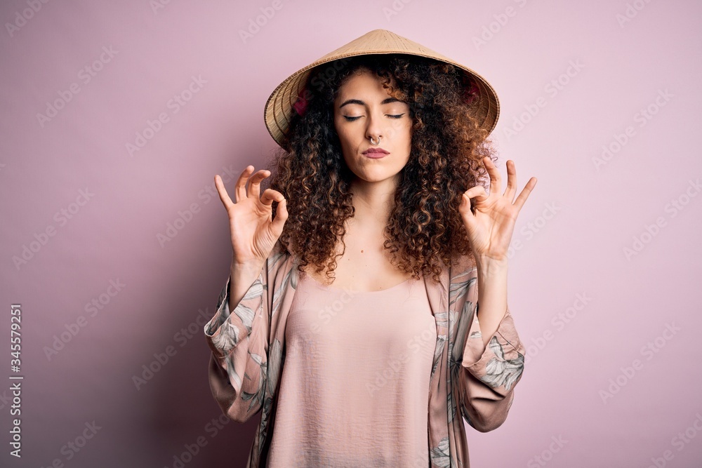 Young beautiful woman with curly hair and piercing wearing traditional asian conical hat relax and smiling with eyes closed doing meditation gesture with fingers. Yoga concept. Young beautiful woman with curly hair and piercing wearing traditional asian conical hat relax and smiling with eyes closed doing meditation gesture with fingers. Yoga concept.