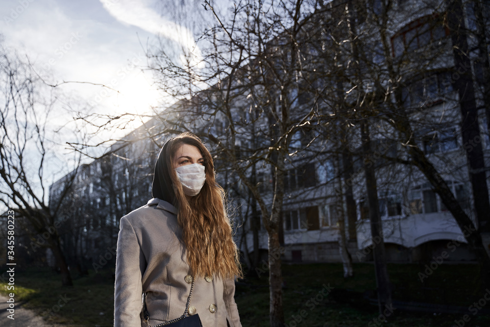 girl with long hair in a protective medical mask stands in the sunlight against the background of trees and houses and looks away girl with long hair in a protective medical mask stands in the sunlight against the background of trees and houses and looks away