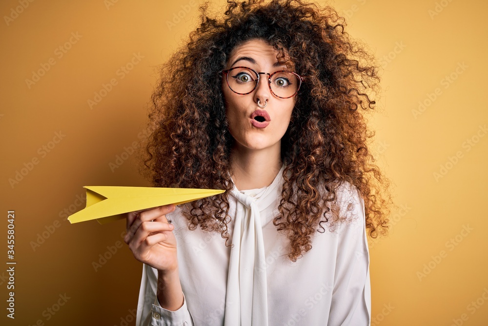 Beautiful woman with curly hair and piercing holding paper plane over yellow background scared in shock with a surprise face, afraid and excited with fear expression Beautiful woman with curly hair and piercing holding paper plane over yellow background scared in shock with a surprise face, afraid and excited with fear expression