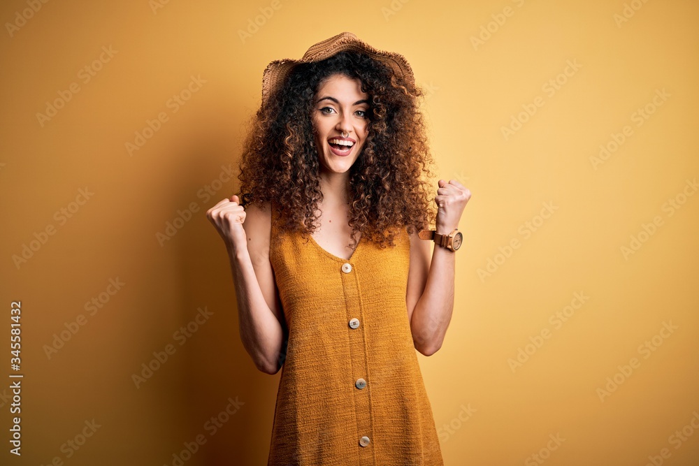 Beautiful brunette woman on vacation with curly hair and piercing wearing hat and dress celebrating surprised and amazed for success with arms raised and open eyes. Winner concept. Beautiful brunette woman on vacation with curly hair and piercing wearing hat and dress celebrating surprised and amazed for success with arms raised and open eyes. Winner concept.