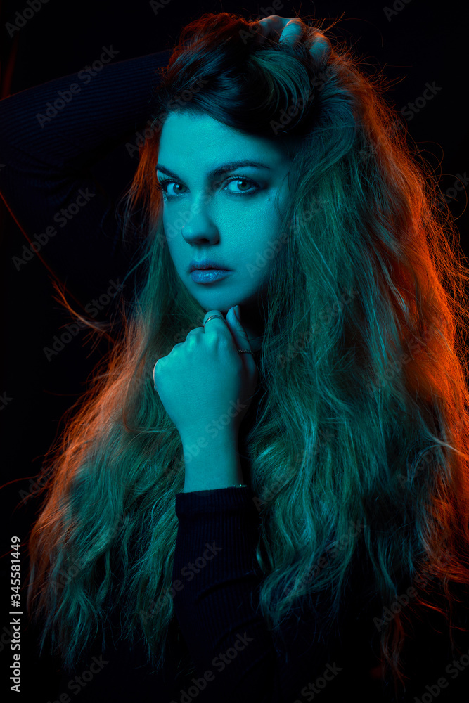 fashion portrait of a blonde girl with color gel filters, neon skin. she holds her hand under her chin, with her second hand holds her hair. blue face, orange hair in the back
