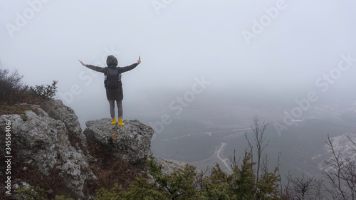 A young woman with a backpack on the edge of a cliff with her hands raised. Misty view of the valley. Crimean mountains in the clouds. Travel to Russia.