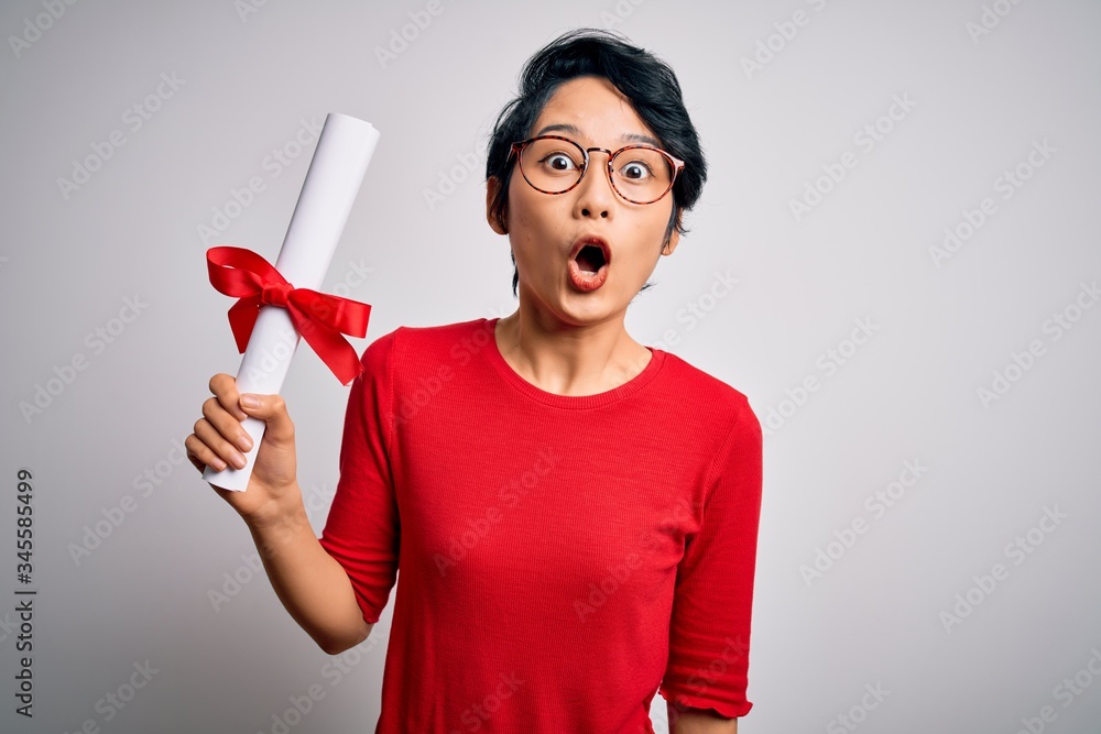 Beautiful chinese student woman wearing glasses holding university graduated diploma degree scared in shock with a surprise face, afraid and excited with fear expression Beautiful chinese student woman wearing glasses holding university graduated diploma degree scared in shock with a surprise face, afraid and excited with fear expression