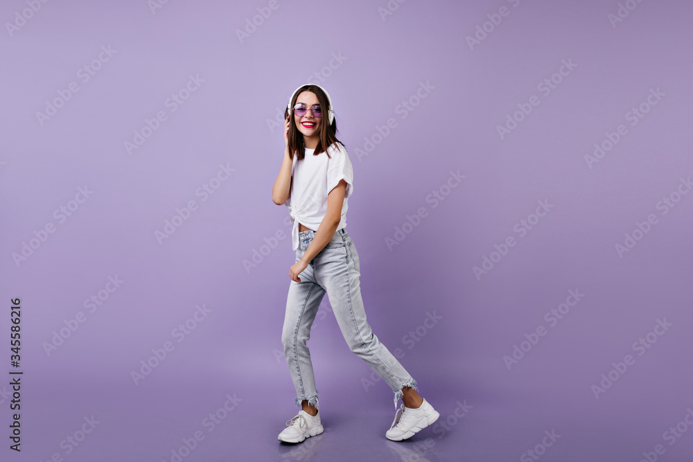 Full-length shot of slim girl in jeans listening music in headphones. Studio portrait of female model in white sneakers dancing on purple background. Full-length shot of slim girl in jeans listening music in headphones. Studio portrait of female model in white sneakers dancing on purple background.