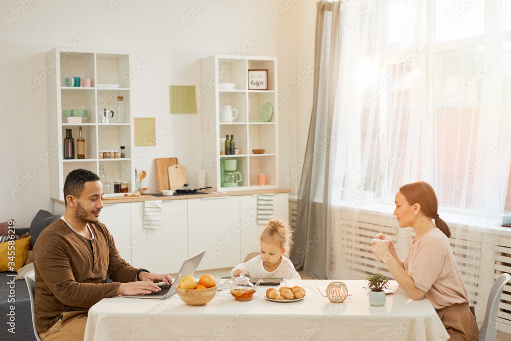 Young man, his wife and little daughter sitting together at table in modern kitchen in morning, horizontal shot