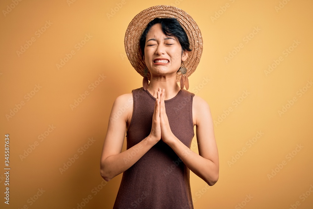 Young beautiful asian girl wearing casual t-shirt and hat over isolated yellow background begging and praying with hands together with hope expression on face very emotional and worried. Begging.