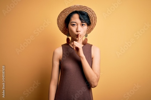 Young beautiful asian girl wearing casual t-shirt and hat over isolated yellow background asking to be quiet with finger on lips. Silence and secret concept.